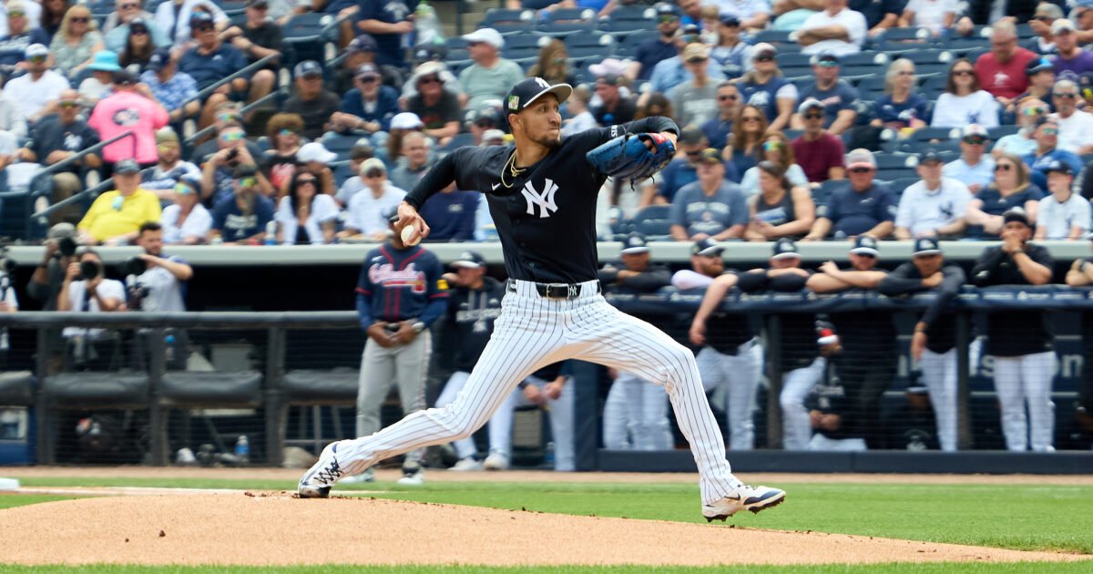New York Yankees pitcher in a black jersey delivering a pitch from the mound with a crowd watching in the stands behind him.