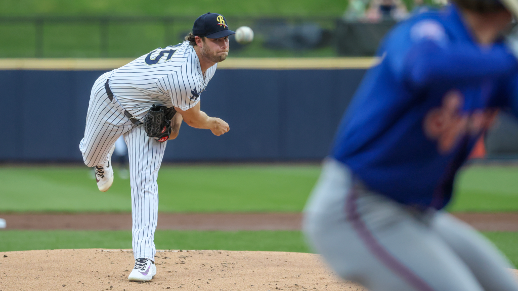 Baseball pitcher in a pinstriped uniform delivers a pitch from the mound, with a blurred batter in blue in the foreground.
